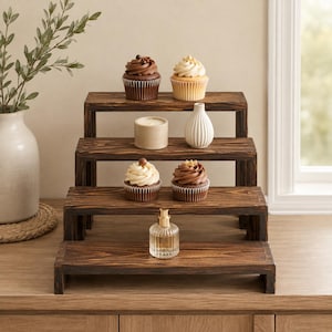 May include: A four-tiered wooden display stand with a dark brown wood grain finish. The stand features rectangular shelves arranged in a cascading staircase design, holding four cupcakes, a cylindrical candle, a small white vase, and a glass bottle.