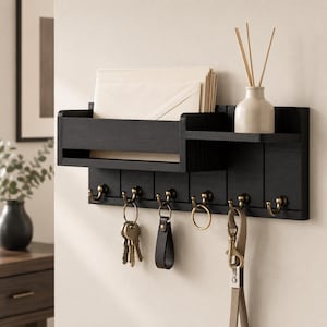 May include: A black wall-mounted organizer featuring a mail slot, a small shelf holding a beige vase with reed sticks, and six brass-toned hooks. Several sets of keys and a lanyard hang from the hooks, while envelopes are stored in the mail slot.