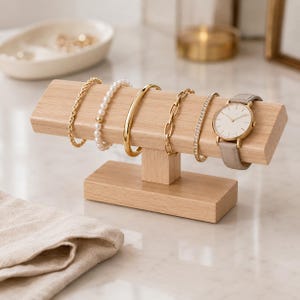 Op de afbeelding: Een T-vormige sieradenstandaard van lichtgekleurd hout met een gouden schakelarmband, een witte parelarmband, een gouden armband, een gouden paperclip-schakelarmband, een dunne armband met kristallen en een polshorloge met een beige leren band.