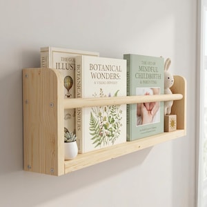 May include: A light-colored wooden wall shelf with two levels, holding books with titles like "Botanical Wonders" and "The Art of Mindful Childbirth & Parenting." A small stuffed bunny and a potted succulent are also on display.