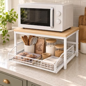 May include: A white microwave sits on a wooden shelf supported by a white metal frame. The lower level features a wire basket containing a cookbook, a white utensil holder with wooden spoons and a whisk, a paper bag, two glass jars, and a folded cloth.