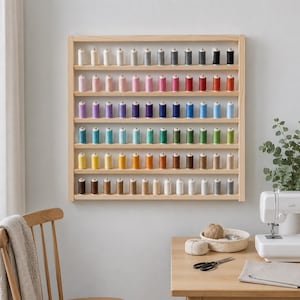 May include: A square light-coloured wooden wall-mounted thread rack featuring six horizontal shelves. Each shelf holds a row of upright sewing thread spools arranged in a gradient of colours, including white, grey, black, pink, red, blue, green, yellow, and brown.