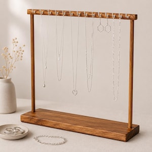May include: A wooden jewelry display stand with a rectangular base and a horizontal bar featuring twelve silver hooks. Several silver necklaces, a pair of hexagonal earrings, a silver bracelet, and two silver rings are displayed on the stand.