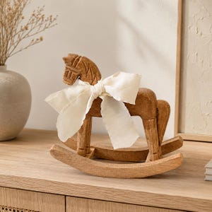 Könnte beinhalten: Eine geschnitzte Schaukelpferd-Figur aus Holz mit einer natürlichen, hellbraunen Holzmaserung. Das Pferd ist mit einer großen, cremeweißen Stoffschleife um den Hals verziert und ruht auf zwei geschwungenen Kufen, die eine sanfte Schaukelbewegung ermöglichen.