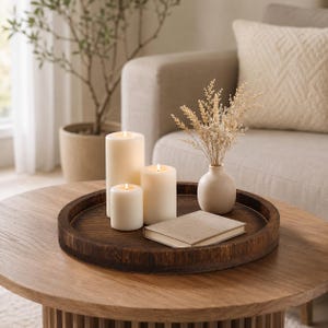 Puede incluir: Una bandeja redonda de madera marrón oscuro que contiene tres velas de pilar de color crema de diferentes alturas, un pequeño jarrón de cerámica beige con hierbas secas claras y un libro rectangular cerrado de tapa lisa.