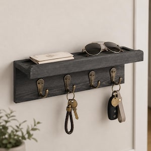 May include: A dark gray wooden wall-mounted shelf featuring four antique brass-toned metal hooks. The shelf holds a folded white paper item and a pair of dark-tinted sunglasses. Two sets of keys with keychains hang from the metal hooks below.
