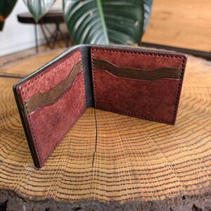 Peut inclure: Portefeuille en cuir bifold ouvert, avec un extérieur foncé et un intérieur bordeaux. Le portefeuille comporte plusieurs fentes pour cartes et un compartiment à billets. Le portefeuille est présenté ouvert sur une surface en bois.