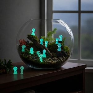 May include: A clear glass bowl terrarium containing green moss, small pebbles, a piece of driftwood, and several small, glowing cyan-coloured humanoid figurines. Two additional glowing figurines are placed outside the bowl on the surface.