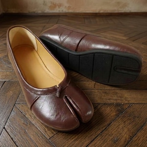 Op de afbeelding: Een paar donkerbruine schoenen met gespleten teen, voorzien van een gladde lederachtige buitenkant, een lage instap en een beige binnenvoering. De schoenen hebben een platte zwarte rubberen zool met een gestructureerd profiel en een teenstuk.