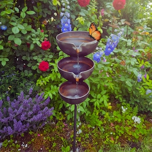 Può includere: Una fontana d'acqua a tre livelli con tre vasche in metallo, ognuna con acqua a cascata nella successiva. Una farfalla arancione si posa sulla vasca superiore. La fontana è in un giardino fiorito.