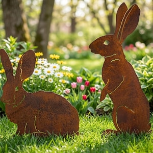 May include: Two rusty brown metal rabbit garden sculptures. One rabbit is sitting, the other is standing upright. The sculptures are placed in a garden with green grass and colourful flowers in the background.