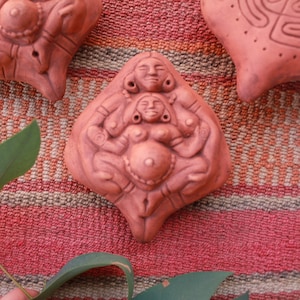 Op de afbeelding: Een terracotta sculptuur die een zwangere vrouw afbeeldt met twee andere figuren, een aan elke kant van haar. Het beeld heeft een diamantvorm en een gedetailleerd ontwerp.