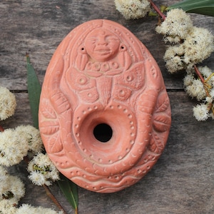Op de afbeelding: Een terracotta olielamp met een gedetailleerd ontwerp van een vrouwengezicht en een slang. De lamp heeft een rond gat in het midden voor de lont.