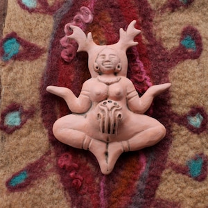 May include: A pink ceramic figurine of a woman with antlers, sitting in a meditative pose with her hands resting on her lap. The figurine is on a brown and red patterned background.