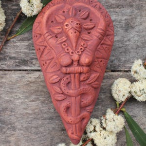 Op de afbeelding: Een wandhanger van terracotta klei in de vorm van een hart met een gedetailleerd ontwerp van een slang die om een ​​staf is gewikkeld. Het ontwerp is roodbruin van kleur.