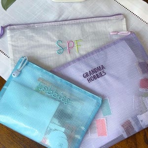 May include: Three rectangular mesh zipper pouches in white, light blue, and lavender. The white pouch features the embroidered text "SPF" in multi-colored thread. The lavender pouch displays "GRANDMA HOBBIES" and the blue pouch says "hobbies".