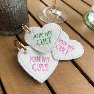 May include: Three white heart-shaped keychains with gold-colored key rings. Each keychain has the embroidered text "JOIN MY CULT" in either green or pink. The keychains are on a wooden surface.