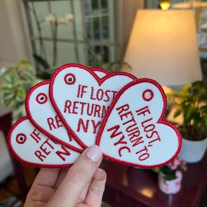 May include: Heart-shaped white patches with red trim and the text "IF LOST RETURN TO NYC". Each patch has a red and white target-like design. The patches are held in a hand.