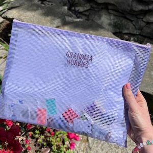 May include: A rectangular, light purple mesh zipper pouch featuring the text "GRANDMA HOBBIES" in dark purple lettering. The semi-transparent pouch contains various small, colorful craft supplies and spools visible through the mesh material.