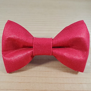 May include: A red bow tie made of fabric with a subtle shimmer. The bow tie is tied and ready to wear.