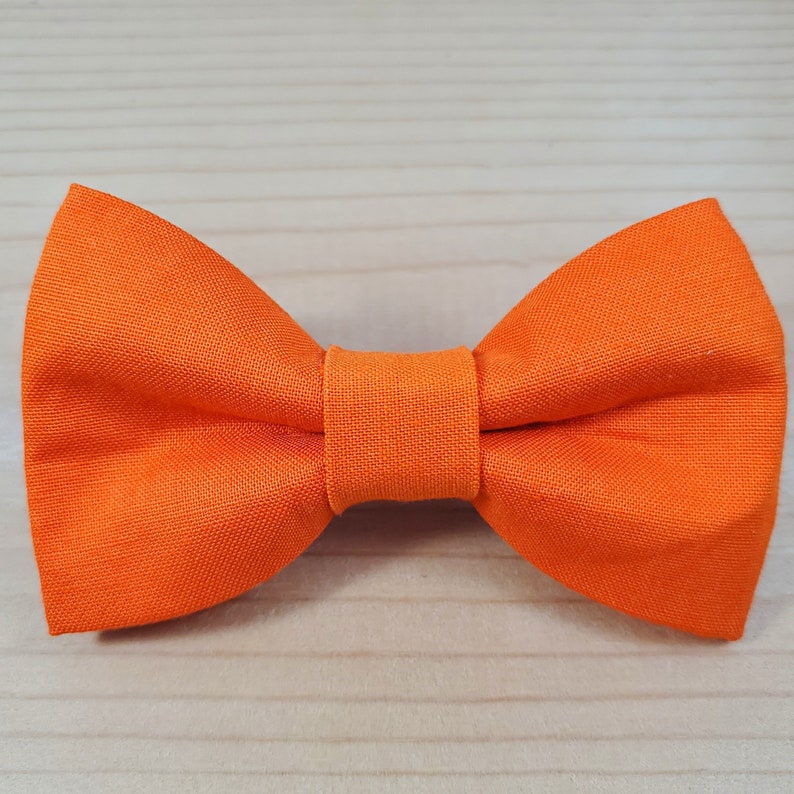 Puede incluir: Una pajarita de color naranja brillante hecha de tela. La pajarita est&aacute; atada y lista para usar.