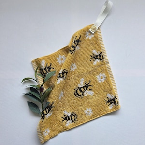 May include: Yellow hand towel with a bee and flower pattern. The towel has a white loop and button for hanging. The edges are trimmed with white and yellow stripes. A sprig of greenery is placed near the towel.