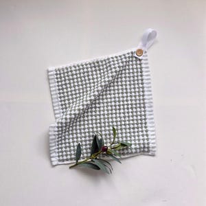 May include: A white and gray patterned kitchen towel with a loop and wooden button for hanging. The towel has a textured design and is accented with a sprig of green leaves and a single dark berry.