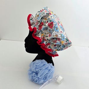 Puede incluir: Un gorro de ducha con un estampado floral multicolor sobre fondo blanco y un borde con volantes de color rojo brillante. El gorro se muestra sobre una cabeza de maniquí negra junto a una esponja de baño de malla azul claro y un cepillo corporal.