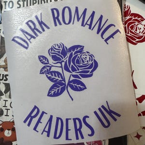 May include: A clear phone case with various stickers. A white sticker with the words "DARK ROMANCE READERS UK" in blue, and a floral design of two roses. Other stickers include the text "I'M ALLERGIC TO STUPIDITY".