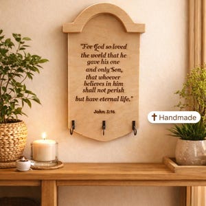 Puede incluir: Portallaves de madera con parte superior curvada, con el texto "Porque de tal manera amó Dios al mundo..." de Juan 3:16. Incluye tres ganchos negros para llaves. Las palabras "A CHERISHED GIFT" están en la parte superior. Una etiqueta "Handmade" está a la derecha.