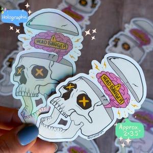 May include: Holographic stickers featuring a skull with an open top, revealing a pink brain labeled "HEAD BANGER." The skull has yellow cross-shaped eyes. The stickers are approximately 2 x 3.5 inches.