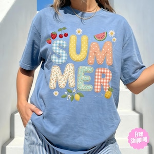 May include: A pale blue t-shirt featuring the word "SUMMER" in a patchwork design. The design incorporates strawberries, cherries, a watermelon slice, daisies, lemons, and a pineapple. The shirt has short sleeves and a loose fit.