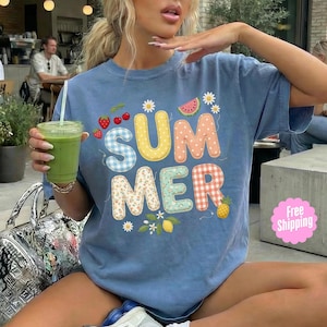 Pode incluir: Camiseta azul com a palavra "SUMMER" em letras coloridas em estilo patchwork. O design inclui morangos, melancia, margaridas, um limão e um abacaxi. Uma peça casual com tema de verão.