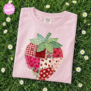 Puede incluir: Una camiseta rosa claro con un gran diseño de fresa en patchwork. La fresa está hecha de varias telas, incluyendo cuadros rojos, estampados florales y patrones de corazones. La camiseta está sobre hierba verde con pequeñas margaritas blancas.