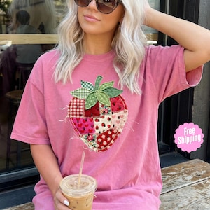 Puede incluir: Una camiseta rosa con un gran diseño de fresa en patchwork. La fresa está hecha de varias telas estampadas, incluyendo corazones, lunares y estampados florales. La camiseta tiene mangas cortas y cuello redondo.