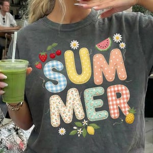 Puede incluir: Camiseta gris oscuro con la palabra "SUMMER" en letras coloridas y estampadas. El diseño incluye fresas, cerezas, una rodaja de sandía, margaritas, un limón y una piña. La camiseta tiene un aspecto vintage.