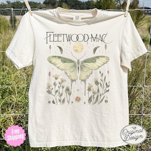 May include: Off-white t-shirt with the text "Fleetwood Mac" above a detailed illustration of a butterfly, moon, and floral elements. The design includes the words "Original Design" in a circular logo.