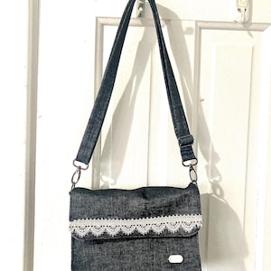Puede incluir: Bolso cruzado de mezclilla oscura con un ribete de encaje blanco en la solapa. El bolso tiene una correa larga y ajustable y herrajes plateados. El bolso está colgado de una puerta blanca.