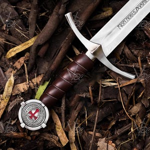 May include: A medieval sword with a polished steel blade and a brown leather-wrapped handle. The blade has the inscription "NON NOBIS, DOMINE". The hilt features a silver crossguard and a decorative pommel with a red cross emblem. The sword rests on a bed of leaves and twigs.