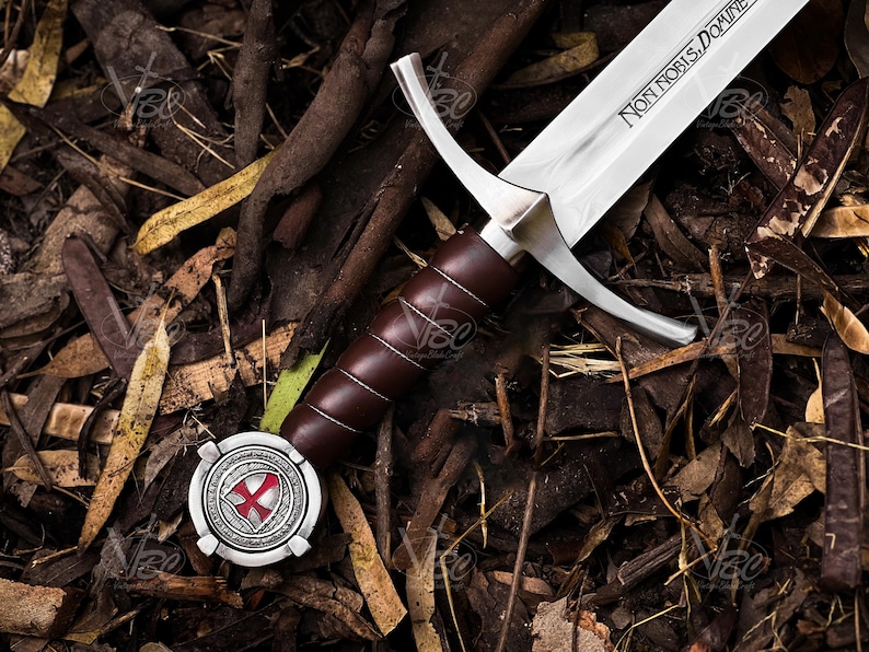 May include: A medieval sword with a polished steel blade and a brown leather-wrapped handle. The blade has the inscription "NON NOBIS, DOMINE". The hilt features a silver crossguard and a decorative pommel with a red cross emblem. The sword rests on a bed of leaves and twigs.