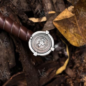 May include: Close-up of a sword hilt with a brown leather-wrapped grip and a silver pommel. The pommel features a detailed design with a central emblem and decorative border. The sword rests on a bed of leaves and wood.