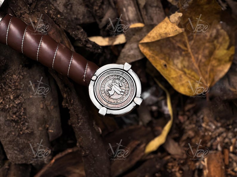 May include: Close-up of a sword hilt with a brown leather-wrapped grip and a silver pommel. The pommel features a detailed design with a central emblem and decorative border. The sword rests on a bed of leaves and wood.