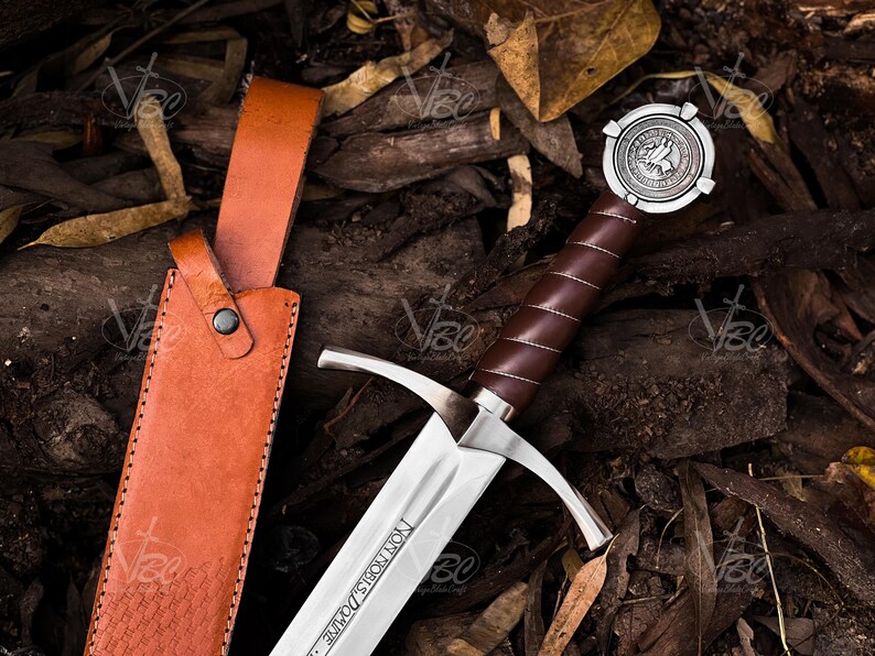 May include: A medieval-style sword with a brown leather-wrapped handle and a silver crossguard. The blade is silver with text etched on it. The sword is next to a brown leather sheath. The sword's pommel has a circular design.