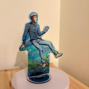 Könnte beinhalten: Ein Acryl-Aufsteller mit einer Anime-Figur mit kurzem braunem Haar, die ein dunkelblaues Hemd und eine hellblaue Jeans trägt. Die Figur sitzt auf einem Baumstamm in einer grasbewachsenen, wasserreichen Umgebung und ist auf einem runden Sockel montiert.