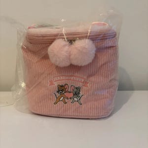 May include: A pink corduroy cosmetic bag with a zipper closure and two fluffy pom-poms. The bag features an embroidered design of Jerry and Tuffy from the cartoon series. The bag is packaged in clear plastic.