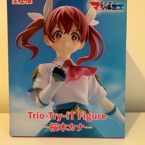 Könnte beinhalten: Verpackte Anime-Figur eines Mädchens mit roten Zöpfen, das eine weiße und blaue Schuluniform trägt. Auf der Box steht "Trio-Try-iT Figure" und japanischer Text. Der Hintergrund ist ein blauer Himmel mit Wolken.
