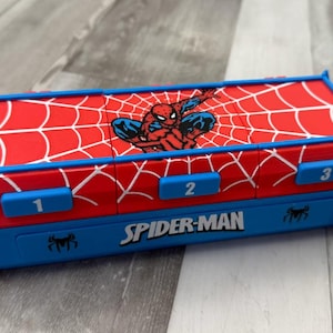 Puede incluir: Un estuche rectangular de plástico rojo y azul con un gráfico de Spider-Man en la tapa. La tapa tiene un patrón de telaraña blanca y tres botones azules numerados 1, 2 y 3. El panel frontal muestra el texto SPIDER-MAN en color blanco.