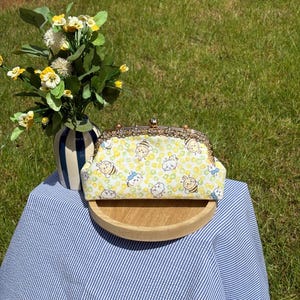 May include: A rectangular fabric clutch purse featuring a repeating pattern of cartoon bees and small white creatures on a yellow and light green background. The purse has a metallic gold-toned kiss-lock frame with decorative floral-like detailing.