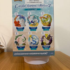 May include: A rectangular product box for the Pokemon Circular Diorama Collection 2. The box features images of six distinct circular dioramas featuring various Pokemon characters, including Vulpix, Alola Vulpix, Primarina, Flygon, Mew, Dragonair, Mimikyu, and Phantump.