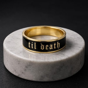 Könnte beinhalten: Ein goldfarbener Ring mit einem schwarzen Band, auf dem in goldener Schrift "til death" steht. Der Ring liegt auf einer runden, marmorähnlichen Oberfläche. Das Design des Rings ist auffällig.